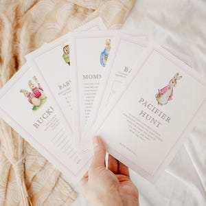 May include: A hand holds a stack of baby shower game cards. Each card features a watercolor illustration of a bunny and has a pink border. The cards include games like "Pacifier Hunt" and "Baby Bingo".