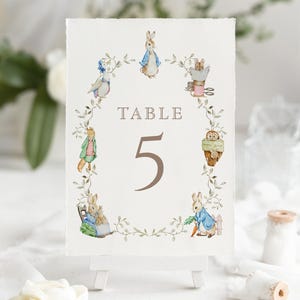 May include: A watercolor table number card featuring illustrations of Peter Rabbit characters. The card displays the word "TABLE" and the number "5" in brown. The illustrations are arranged around the text and number, with a leafy border.