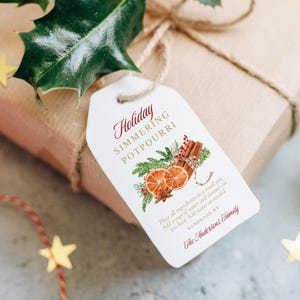 May include: A white gift tag with a red and gold design. The tag says "Holiday Simmering Potpourri" and includes instructions for making the potpourri. The tag is tied with twine to a brown gift box.