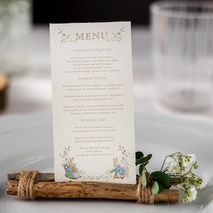 Peut inclure: Une carte de menu verticale de couleur crème avec le texte MENU, une liste de plats et des illustrations de lapins. La carte est maintenue droite par un support rustique composé de deux bâtons de bois horizontaux entourés de ficelle et de verdure.