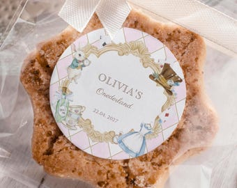 May include: A star-shaped cookie in a clear cellophane bag, tied with a white ribbon. The cookie has a round label with the text "OLIVIA'S Onerderland" and the date "22.04.2027". The label features illustrations from Alice in Wonderland.