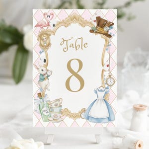 Puede incluir: Una tarjeta decorativa con número de mesa con el tema de Alicia en el país de las maravillas. La tarjeta tiene un marco dorado con ilustraciones de un flamenco, el sombrero del Sombrerero Loco y el vestido de Alicia. El número "8" es dorado.
