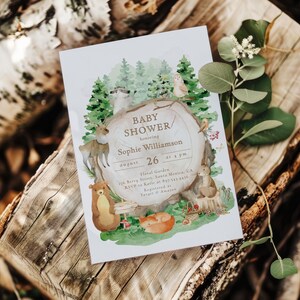 Puede incluir: Una invitación a un baby shower con un tema de bosque, con ilustraciones de animales y un tronco de árbol. La invitación incluye el texto "Baby Shower" y los detalles del baby shower de Sophie Williamson el 26 de agosto a las 4 pm. La invitación está sobre una superficie de madera.