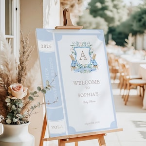 Op de afbeelding: Een welkomstbord voor een babyshower op een houten ezel. Het bord is ontworpen als een boek, met een lichtblauwe rand en de tekst "Welcome to Sophia's Baby Shower". Het heeft een bloemenwapen met twee konijnen.