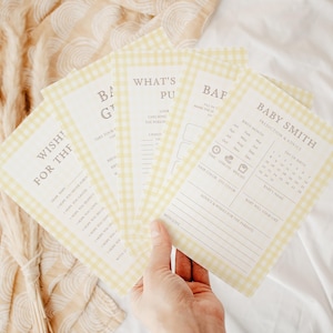 May include: A hand holds a stack of baby shower game cards. The cards have a yellow gingham border and include text such as "Baby Smith" and "Wishes for the Baby". The cards are on a white surface.