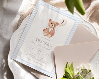 May include: A blue and white striped invitation with a brown teddy bear holding a balloon with the number one on it. The invitation says "Oliver's Bear-y First Birthday" with the date and time of the party. The invitation is on a white plate with a pink envelope and white flowers.