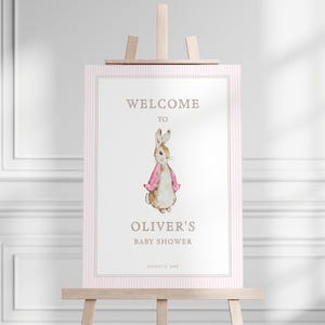 May include: A pink and white striped welcome sign with a watercolor illustration of a rabbit wearing a pink jacket. The sign says "Welcome to Oliver's Baby Shower, August 12, 2023".