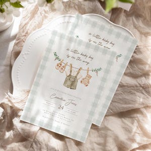 May include: A baby shower invitation with a green and white gingham pattern. The invitation features a watercolour illustration of a baby's clothes hanging on a clothesline with a teddy bear. The text reads "a little baby boy is on the way" and includes the details of the shower, including the date, time, and location.
