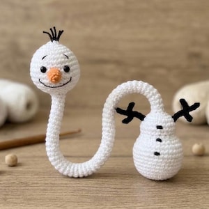May include: A white crocheted snowman figure with a long, curved neck, a small orange carrot nose, black embroidered eyes and mouth, three black buttons on the body, and two black twig-like arms extending from the sides.