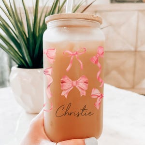 May include: A frosted glass jar with a bamboo lid and a straw. The jar is decorated with pink bows and the name "Christie" is written on the front. The jar is filled with a light brown liquid.