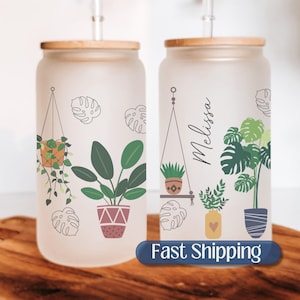 May include: Two frosted glass jars with bamboo lids and a straw. The jars have a white background with a design of hanging plants and potted plants. The jar on the right has the name "Melissa" written on it. The jars are on a wooden surface. Fast Shipping.
