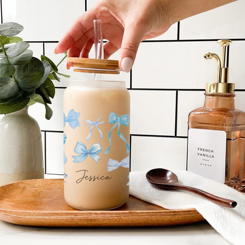May include: A personalized glass jar with a bamboo lid and a straw. The jar has a light brown liquid inside and a blue watercolor bow design. The name "Jessica" is written on the jar.