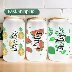 May include: Three white reusable glass tumblers with bamboo lids and straws. The tumblers feature different fruit designs: pineapples, watermelons, and limes. Each tumbler has a name on it: Jessica, Emily, and Caitlin.