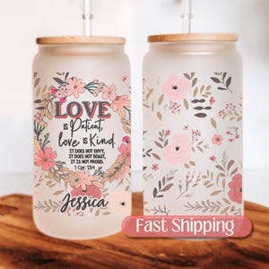 May include: A frosted glass tumbler with a bamboo lid and a straw. The tumbler features a floral design with pink flowers and leaves. The tumbler has a quote about love, "Love is patient, love is kind." The quote is surrounded by a wreath of flowers. The tumbler is personalized with the name "Jessica".