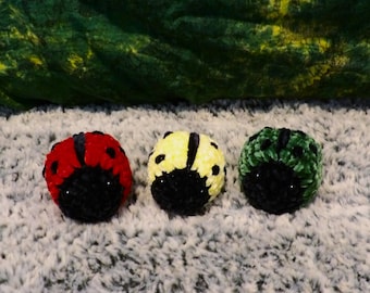 May include: Three plush ladybug toys, one red, one yellow, and one green, with black spots and black faces. The toys are sitting on a gray fuzzy surface.