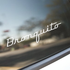 以下が含まれることがあります： 白い筆記体フォントで「Bronquito」という文字が書かれた車の窓のクローズアップ。テキストはデカールまたはステッカーのようで、少し使い古されたように見えます。背景はぼやけており、屋外環境を示唆しています。