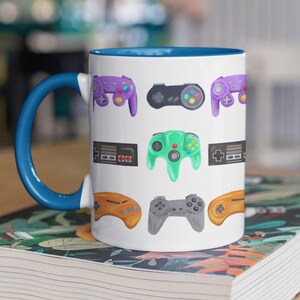 May include: White ceramic coffee mug with a blue rim and handle. The mug features a pattern of colorful video game controllers and game consoles.