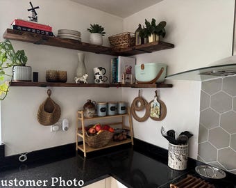 May include: A kitchen with wooden shelves, a black countertop, and a white tiled backsplash. The shelves are filled with various items, including plants, books, and kitchenware. There is a small wooden shelf unit with jars and a basket of fruit. The countertop has a black and white utensil holder and a gray and white tile backsplash.