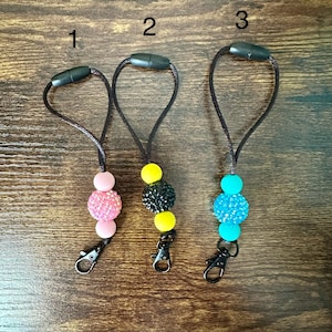 May include: Three black beaded lanyards with a metal clip on one end and a black plastic clasp on the other. Each lanyard has a single, large, colorful, sparkly bead in the center. The beads are pink, yellow, and blue.