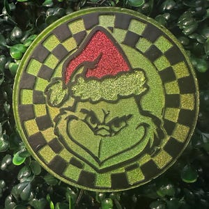 May include: A round, green and black Christmas ornament featuring a Grinch design. The Grinch is wearing a red glitter Santa hat. The ornament has a checkered border and is set against a background of green leaves.