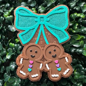May include: A brown gingerbread cookie ornament with a teal bow and two gingerbread people holding hands. The gingerbread people have white icing details and colorful heart accents. The ornament is set against a green background.