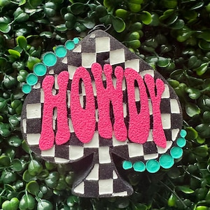 May include: A spade-shaped ornament featuring a black and white checkered pattern. The word HOWDY is written in large, textured, bright pink letters across the center. A series of small, round, turquoise-colored beads line the upper left and lower right edges.