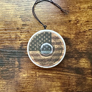 May include: A round wooden ornament with a distressed finish, featuring an American flag design. The ornament has a black cord for hanging.