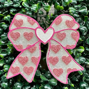 May include: A decorative bow ornament featuring a translucent white base with a textured surface. The bow is adorned with numerous small, pink glittery heart shapes and is outlined in a vibrant pink border, with a central white heart accent.