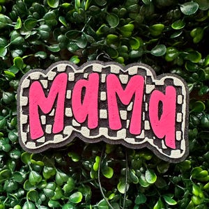 May include: A pink "MAMA" sign with a black and white checkered border. The sign is set against a backdrop of green foliage. The letters are in a bold, bubble-style font.