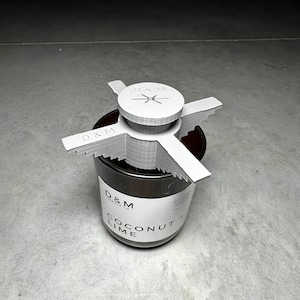 Puede incluir: Tapa de vela blanca con un diseño de cruz y el texto "D&M Candle Co." en la parte superior de un tarro de vela negro con el texto "D&M Candle Co. Coconut Lime" en la etiqueta.