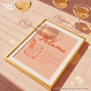K&ouml;nnte beinhalten: Ein goldgerahmter Druck mit rosa Hintergrund zeigt ein Cocktailrezept "Classic Paloma". Das Rezept enth&auml;lt Tequila, Grapefruitsaft, Limettensaft, Grapefruitsoda, Salz und Eisw&uuml;rfel. Drei Gl&auml;ser eines goldenen Getr&auml;nks sind im Hintergrund.