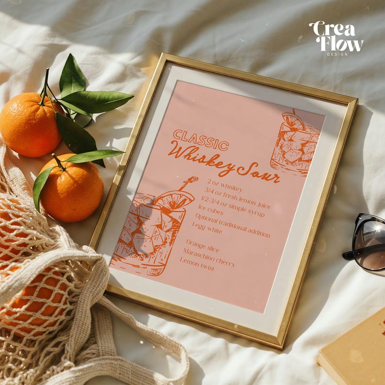 K&ouml;nnte beinhalten: Ein gerahmter Druck mit goldenem Rahmen zeigt ein Rezept f&uuml;r "Classic Whiskey Sour" auf rosa Hintergrund. Orangen und gr&uuml;ne Bl&auml;tter befinden sich neben einer Netztasche mit Orangen. Sonnenbrille und ein Buch sind ebenfalls im Bild.