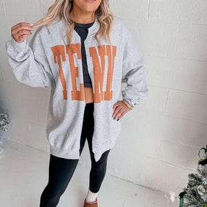 May include: A gray sweatshirt with the word "TENW" in orange block letters. The sweatshirt is open in the front and worn with black leggings and brown slippers. The person is wearing a black top underneath.