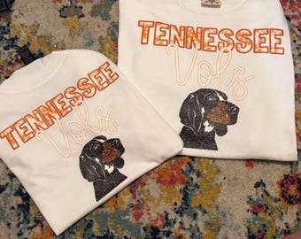 May include: Two white t-shirts with the words "Tennessee Vols" in orange and white lettering. Each shirt features a black and brown dog graphic. The shirts are on a colorful patterned surface.