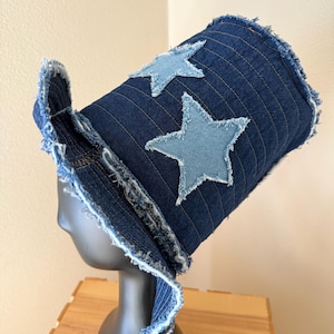 May include: A dark blue denim top hat featuring a frayed brim and edges. The hat is decorated with two light blue denim star-shaped patches stitched onto the side. The fabric has visible vertical stitching lines throughout the cylindrical crown.