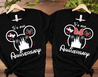 May include: Two black t-shirts with a white Mickey Mouse head design. The shirts say "It's our Anniversary" with a white castle and fireworks inside the Mickey head. One shirt has a red bow and the other has two hands holding a heart.