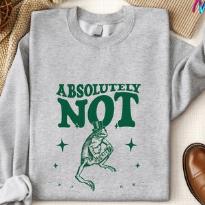 Puede incluir: Una sudadera gris con el texto "ABSOLUTELY NOT" en letras verdes. Debajo del texto hay una rana de dibujos animados con un sombrero de copa y un bast&oacute;n. Hay estrellas verdes alrededor de la rana.
