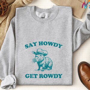 K&ouml;nnte beinhalten: Ein graues Sweatshirt mit dem Text "Say Howdy Get Rowdy" in blaugr&uuml;nen Buchstaben. Ein Cartoon-Capybara mit Cowboyhut befindet sich in der Mitte des Textes.