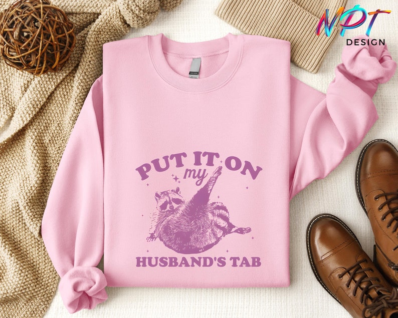 May include: Pink sweatshirt with a purple raccoon graphic and the text "PUT IT ON MY HUSBAND'S TAB".