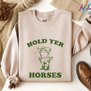 May include: A beige sweatshirt with a green frog wearing a hat and overalls. The text "Hold Yer Horses" is printed in green on the sweatshirt.
