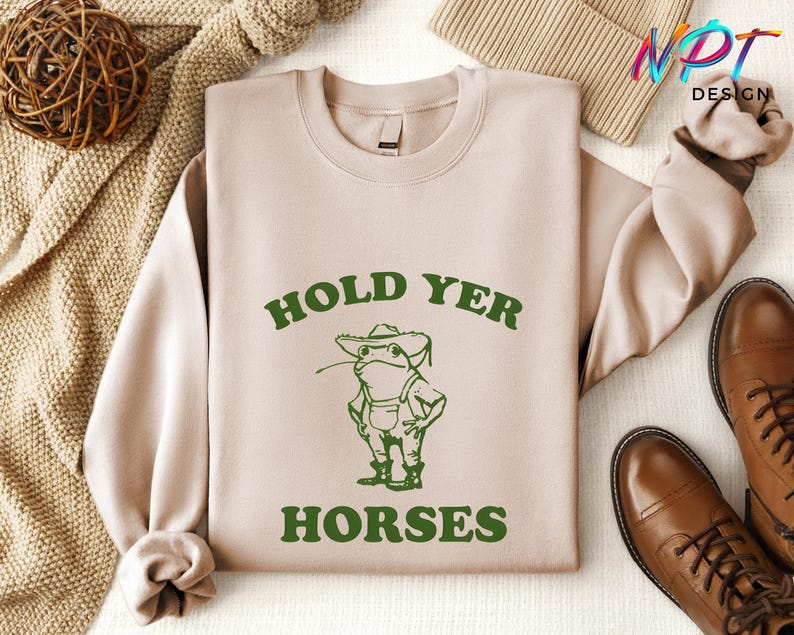 May include: A beige sweatshirt with a green frog wearing a hat and overalls. The text "Hold Yer Horses" is printed in green on the sweatshirt.