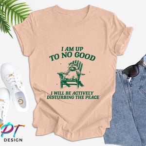 May include: A peach-coloured t-shirt with a green raccoon graphic sitting in a chair. The text "I am up to no good" and "I will be actively disturbing the peace" is printed on the shirt.