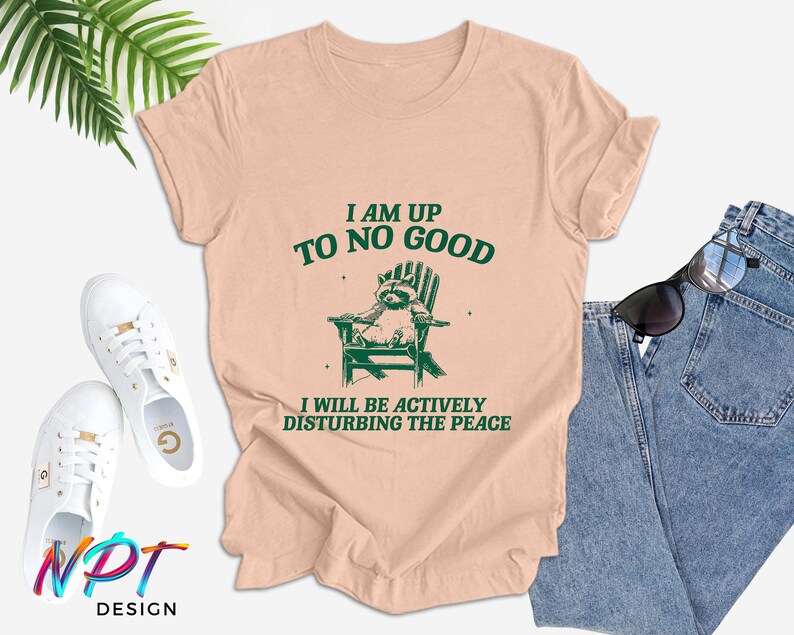 May include: A peach-coloured t-shirt with a green raccoon graphic sitting in a chair. The text "I am up to no good" and "I will be actively disturbing the peace" is printed on the shirt.