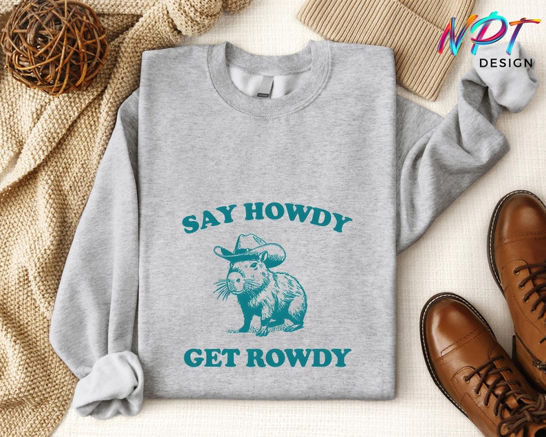 K&ouml;nnte beinhalten: Ein graues Sweatshirt mit dem Text "Say Howdy Get Rowdy" in blaugr&uuml;nen Buchstaben. Ein Cartoon-Capybara mit Cowboyhut befindet sich in der Mitte des Textes.