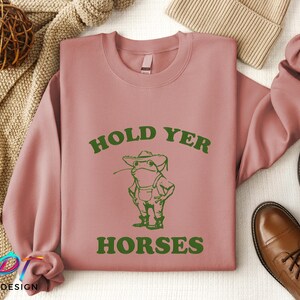 May include: A pink sweatshirt with the text "Hold Yer Horses" in green letters. A cartoon frog wearing a hat and overalls is below the text.