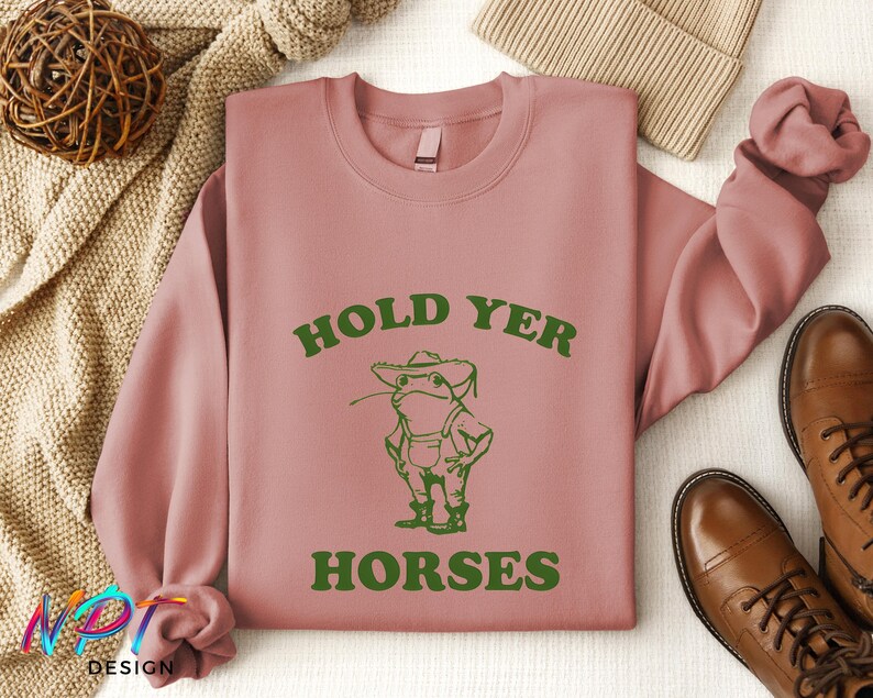 May include: A pink sweatshirt with the text "Hold Yer Horses" in green letters. A cartoon frog wearing a hat and overalls is below the text.