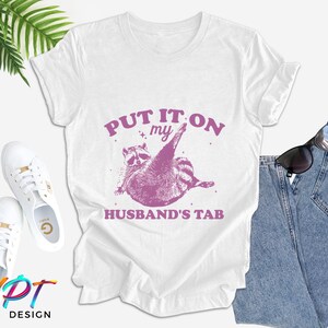 May include: White t-shirt with a pink raccoon graphic and the text "Put it on my husband's tab".