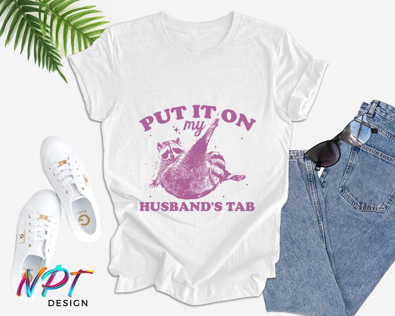 May include: White t-shirt with a pink raccoon graphic and the text "Put it on my husband's tab".