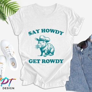 K&ouml;nnte beinhalten: Wei&szlig;es T-Shirt mit einer blaugr&uuml;nen Grafik eines Capybaras, das einen Cowboyhut tr&auml;gt. Der Text "Say Howdy Get Rowdy" ist in blaugr&uuml;n unter dem Capybara gedruckt.