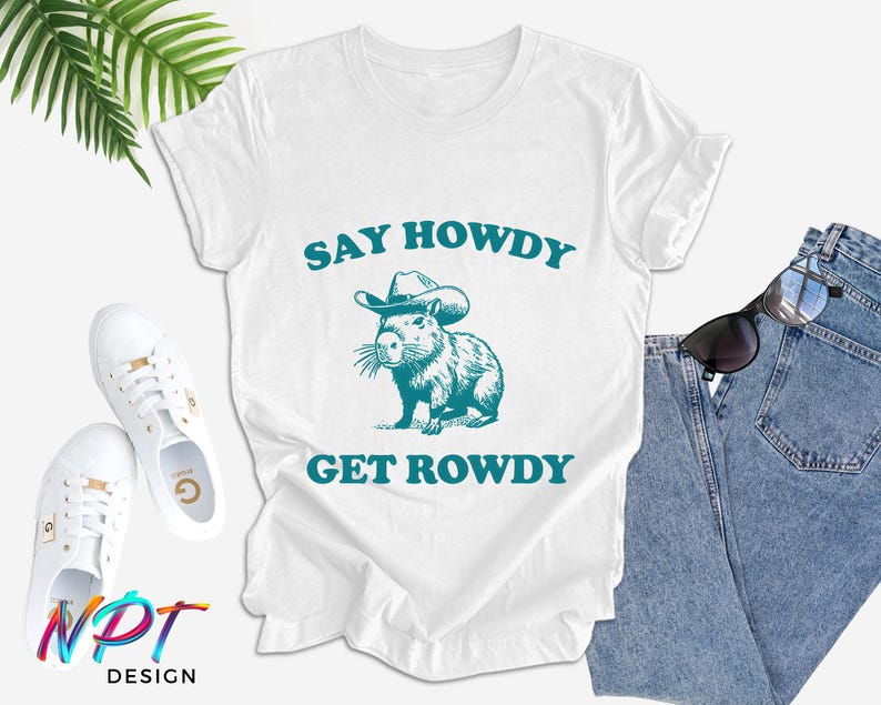 K&ouml;nnte beinhalten: Wei&szlig;es T-Shirt mit einer blaugr&uuml;nen Grafik eines Capybaras, das einen Cowboyhut tr&auml;gt. Der Text "Say Howdy Get Rowdy" ist in blaugr&uuml;n unter dem Capybara gedruckt.